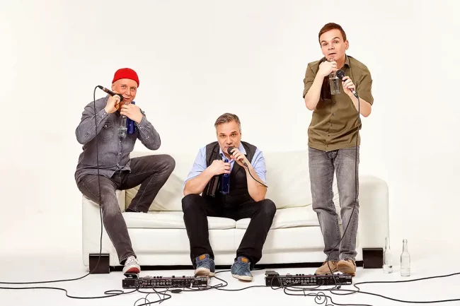 Die drei Musiker der Band GlasBLasSing auf einer Couch mit Instrumenten.