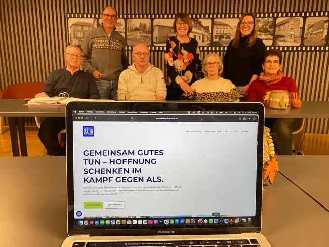 Laptopbildschirm zeigt Internetseite der Gernot-Bohn-ALS-Stiftung, dahinter Mitglieder des Vereins.