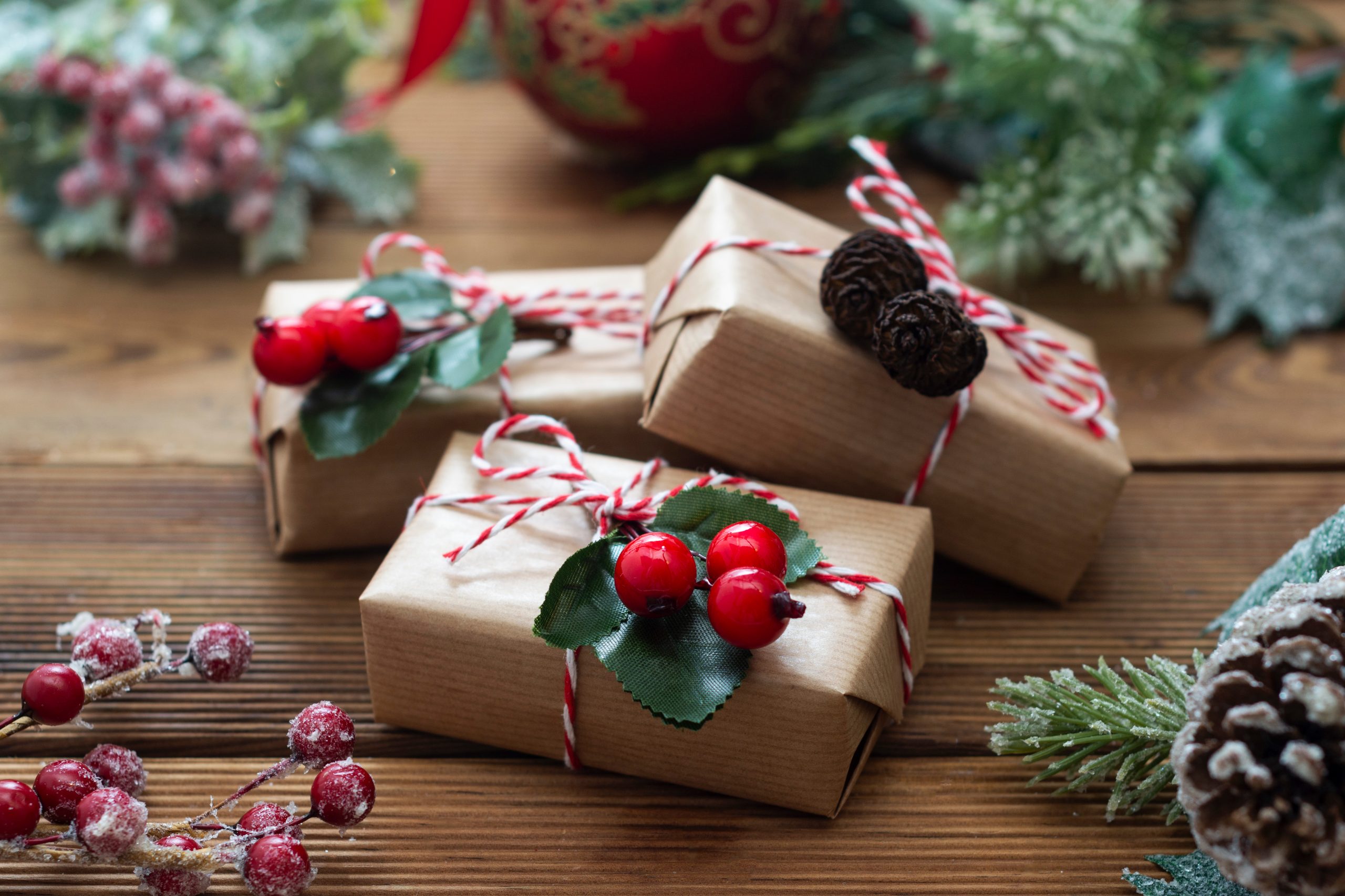 Close up Christmas gift boxes wraped in craft paper, fir branches, red baubles, pine cones over wooden table prepared for celebrating festive holiday season. Weihnachtsgeschenk AdobeStock, Inga